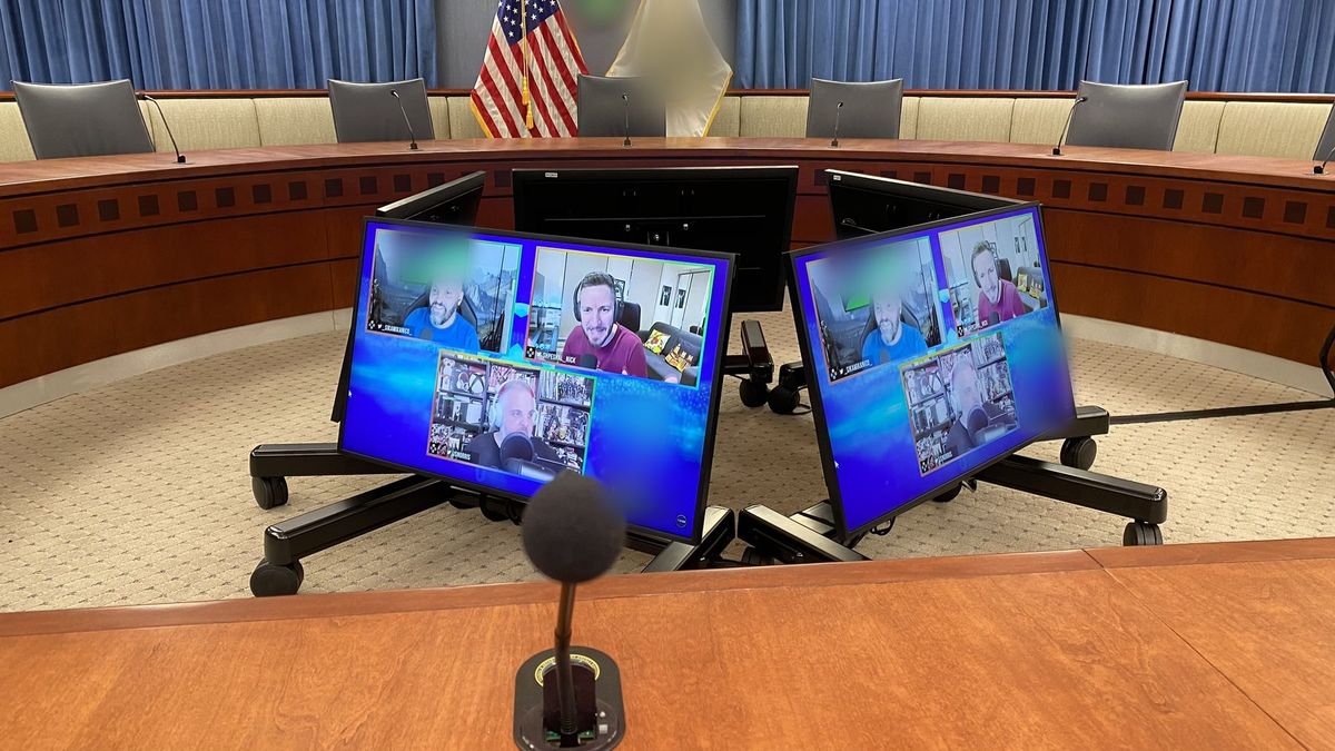 Hearing room dais with gooseneck microphones and motorized monitors showing a live video call, by Enlightened Audiovisual